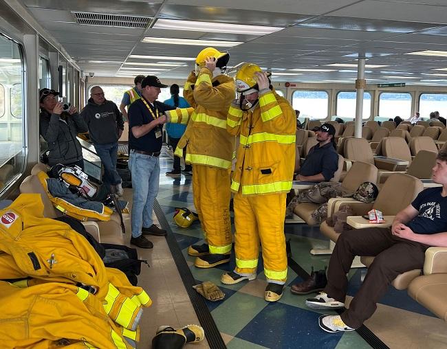 A few people putting on firefighting gear with some helping them and others looking on in the passenger cabin of a ferry